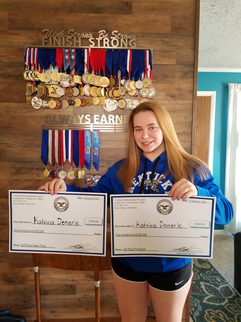Katrina displays medals won in the past 3 years and checks won at the 2020 Camp Perry Open (1st Place 3-Position and 3rd Place Superfinals).